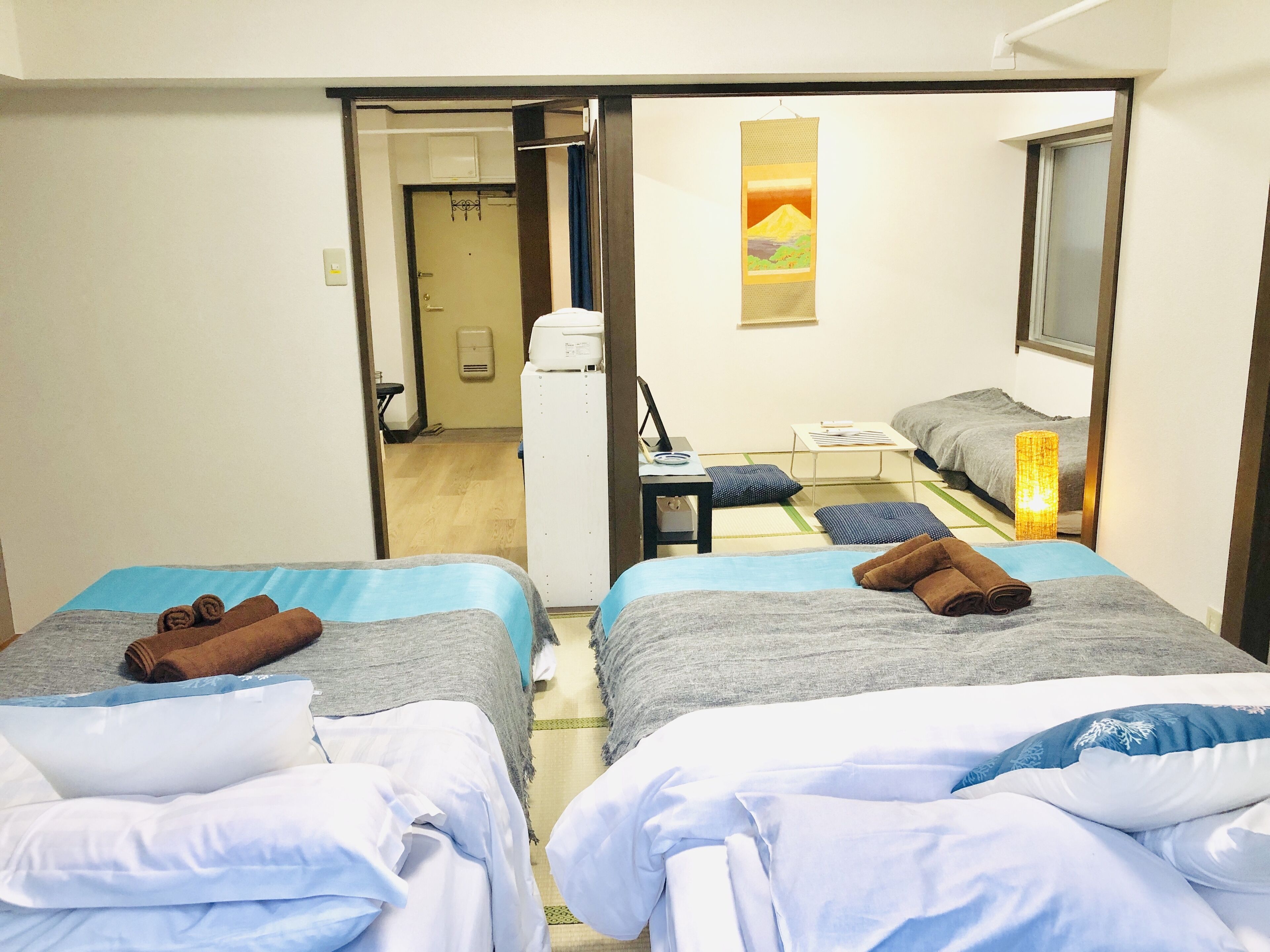 A hotel room with two beds, each with a blue and white bedspread, and brown towels. There is a small desk with a lamp, a wall-mounted air conditioner, and a painting of a mountain.