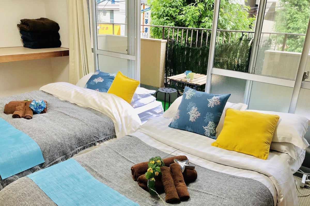 A hotel room with two beds, each with a grey bedspread, blue and yellow pillows, and brown towels. There is a window with a view of greenery outside.