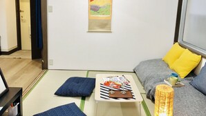 A room with a tatami mat floor, a small wooden table with two glasses, a sofa with yellow cushions, and a wall hanging depicting a mountain. - Rc Annex 2 (Tokyo)