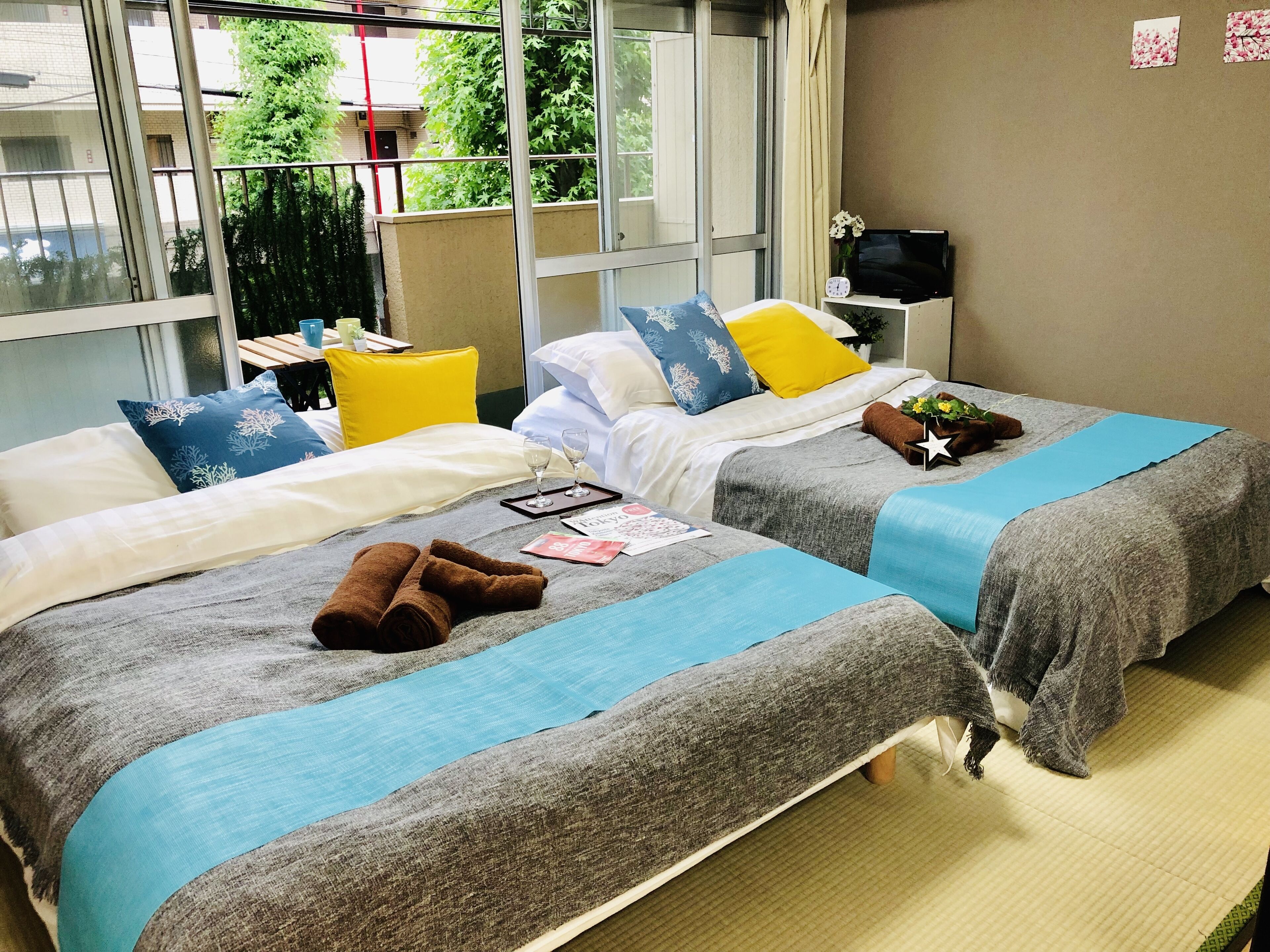 Ein Hotelzimmer mit zwei Betten, jeweils mit blauer und grauer Decke, Kissen und Handtüchern. Ein kleiner Tisch mit einem Tablett, Gläsern und einer Speisekarte steht bereit.