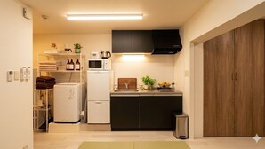 A compact kitchen with a microwave, stove, and sink. There is a small trash bin and a shelf with towels and toiletries. - Rc Annex 2 (Tokyo)