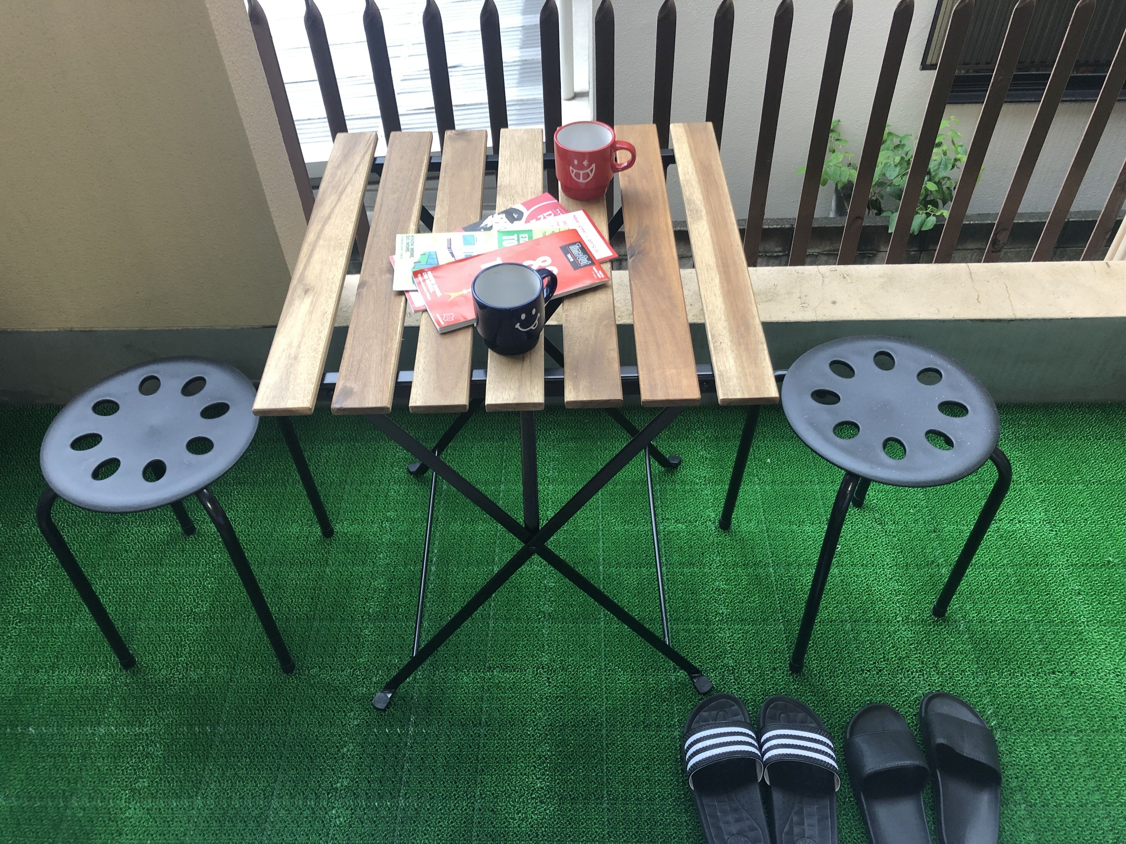 A small outdoor seating area with a wooden table, two black stools, a red mug, and a pair of black slippers on green artificial grass.