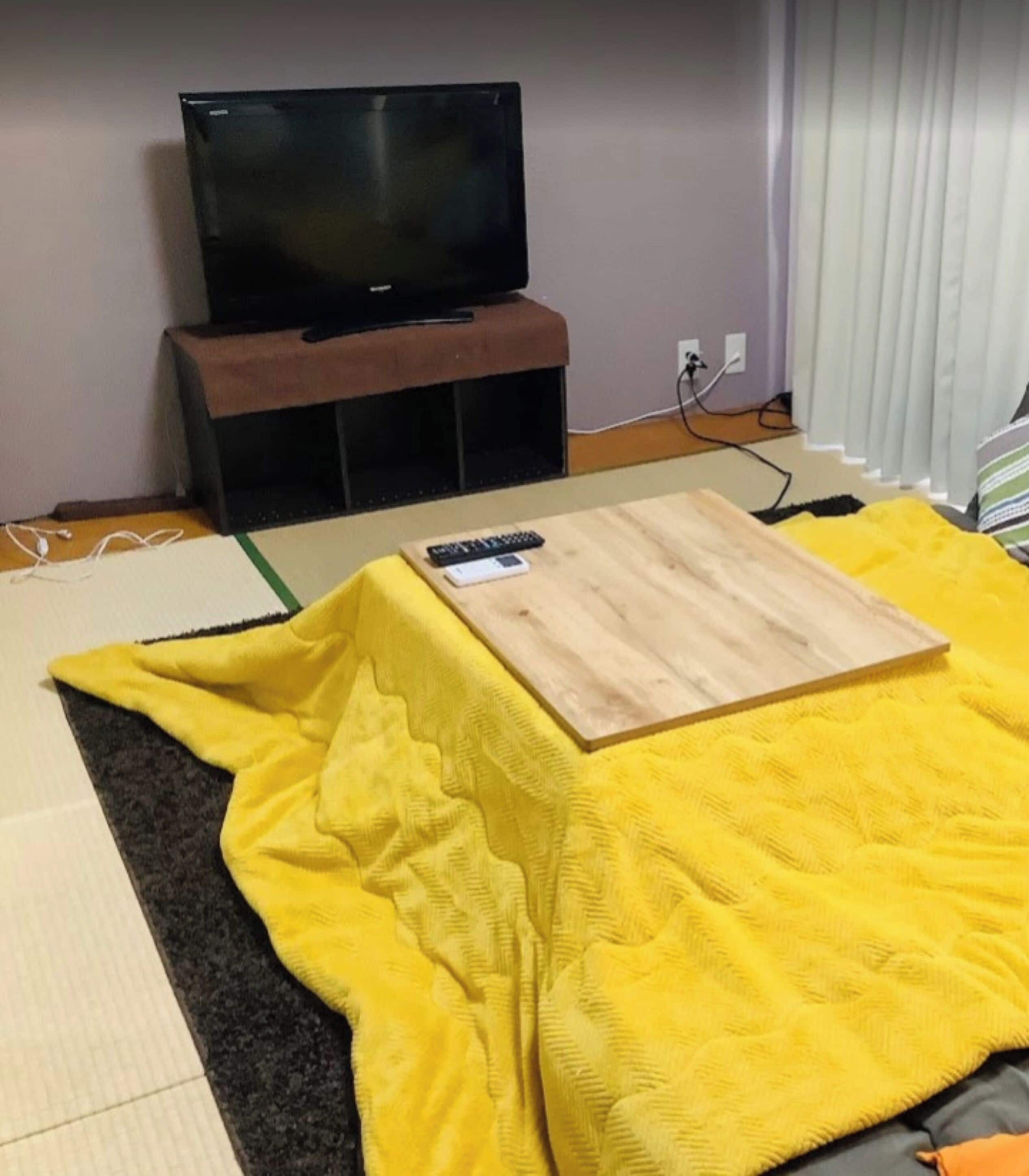 A room with a television on a stand, a wooden table, and a yellow blanket on the floor.