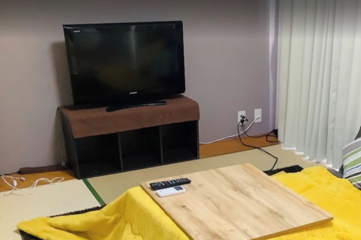 Una habitación con televisión sobre un mueble bajo, una mesa de madera y una manta amarilla en el suelo.