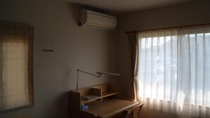 Ett rum med ett skrivbord, en stol, en lampa, ett fönster med tunna gardiner och en luftkonditioneringsanläggning.