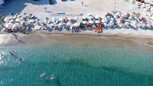 Vista aérea de una playa con agua turquesa cristalina, gente nadando y numerosas sombrillas y sillas de playa.