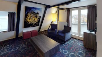 Habitación de hotel con un ventanal grande, un cuadro, un sofá, una silla y una mesa de madera.