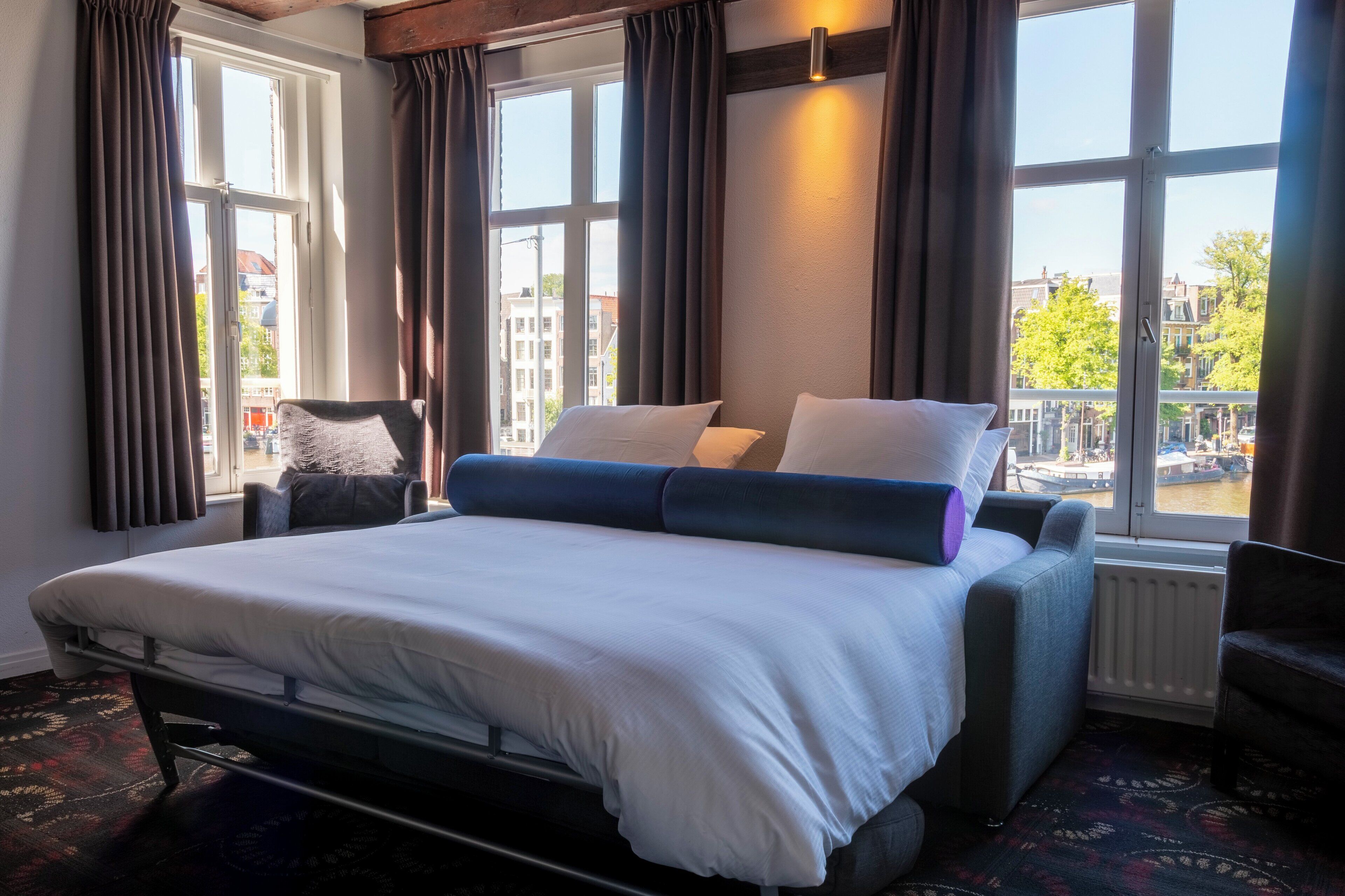 A hotel room with a bed, a chair, a window with curtains, and a view of buildings and a canal.
