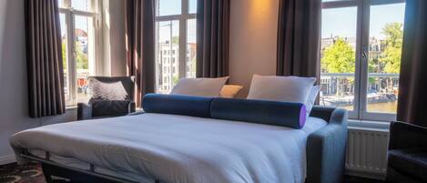 A hotel room with a bed, a chair, a window with curtains, and a view of buildings and a canal.