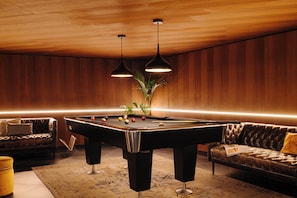 Una mesa de billar en una sala con paredes de madera, un sofá y dos lámparas colgantes.