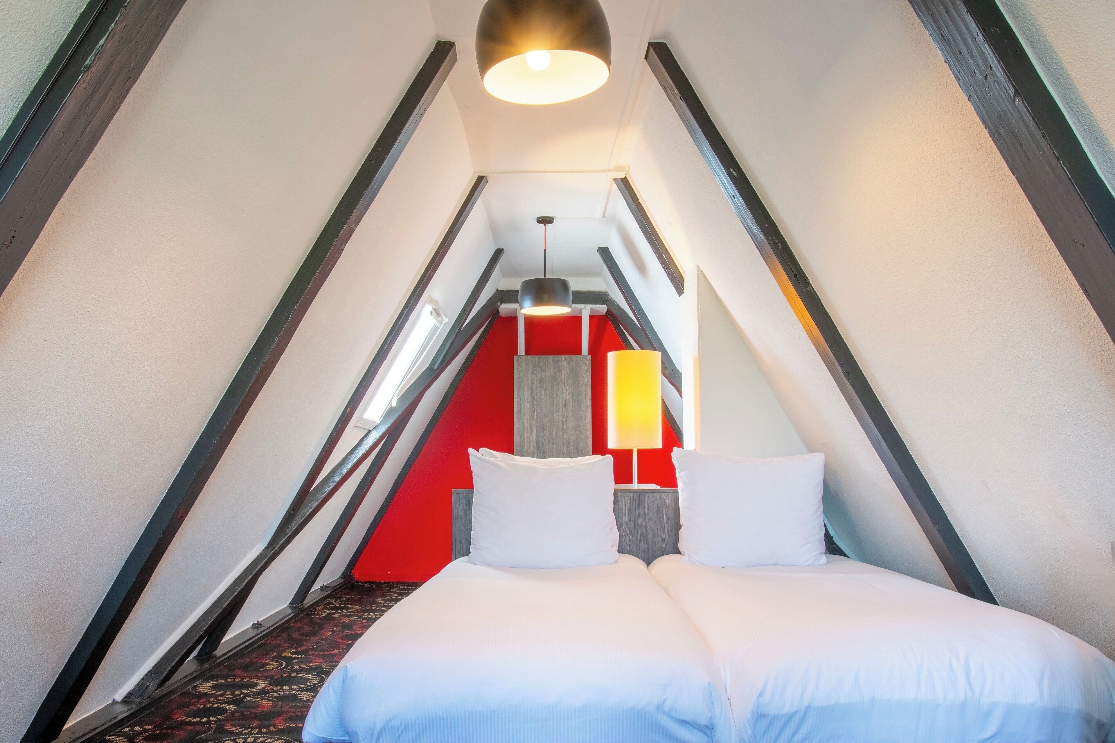 A triangular room with a bed, a red accent wall, and a hanging light fixture.