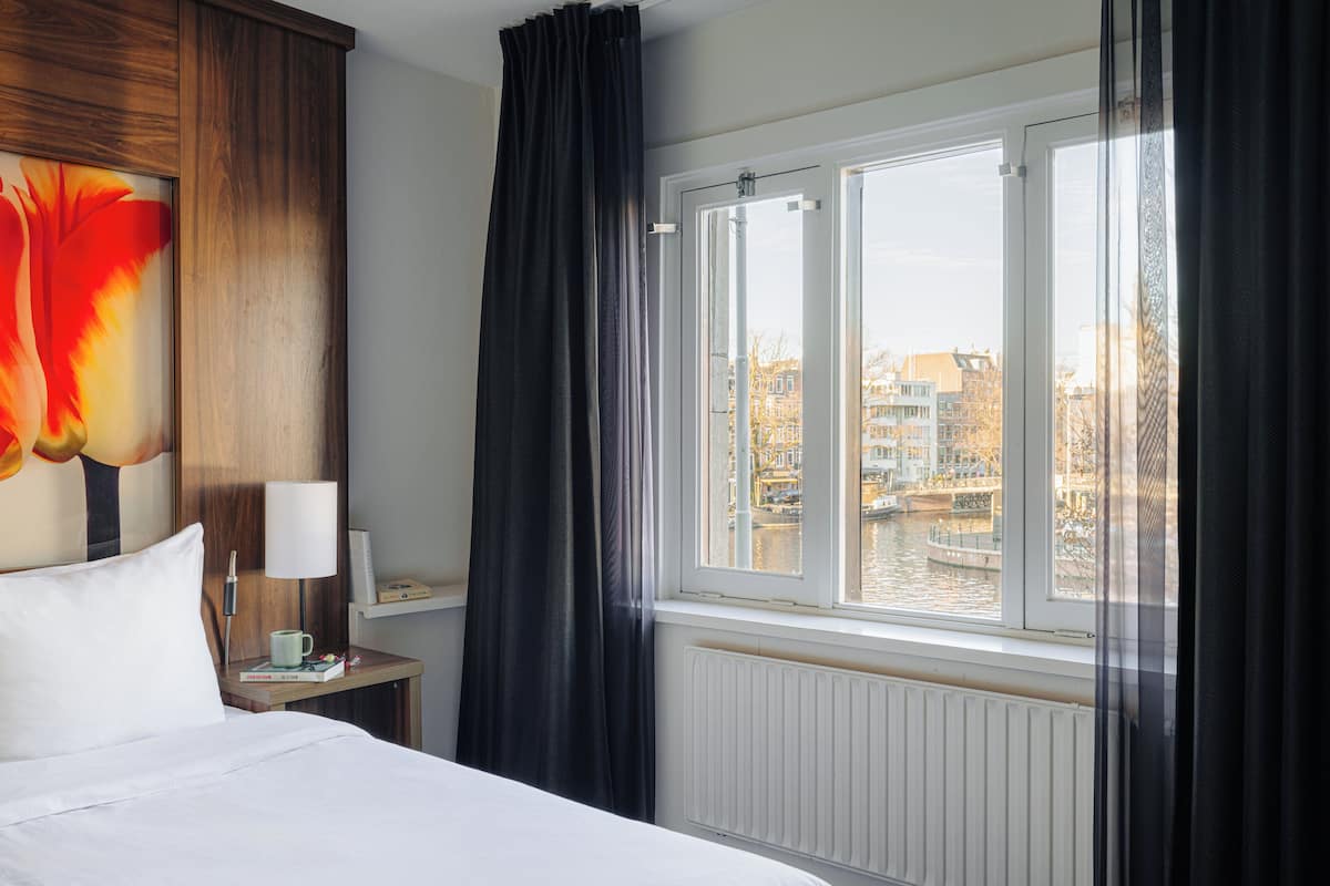 Una habitación de hotel con cama, mesita de noche con lámpara, ventana con vistas a edificios y un cuadro en la pared.