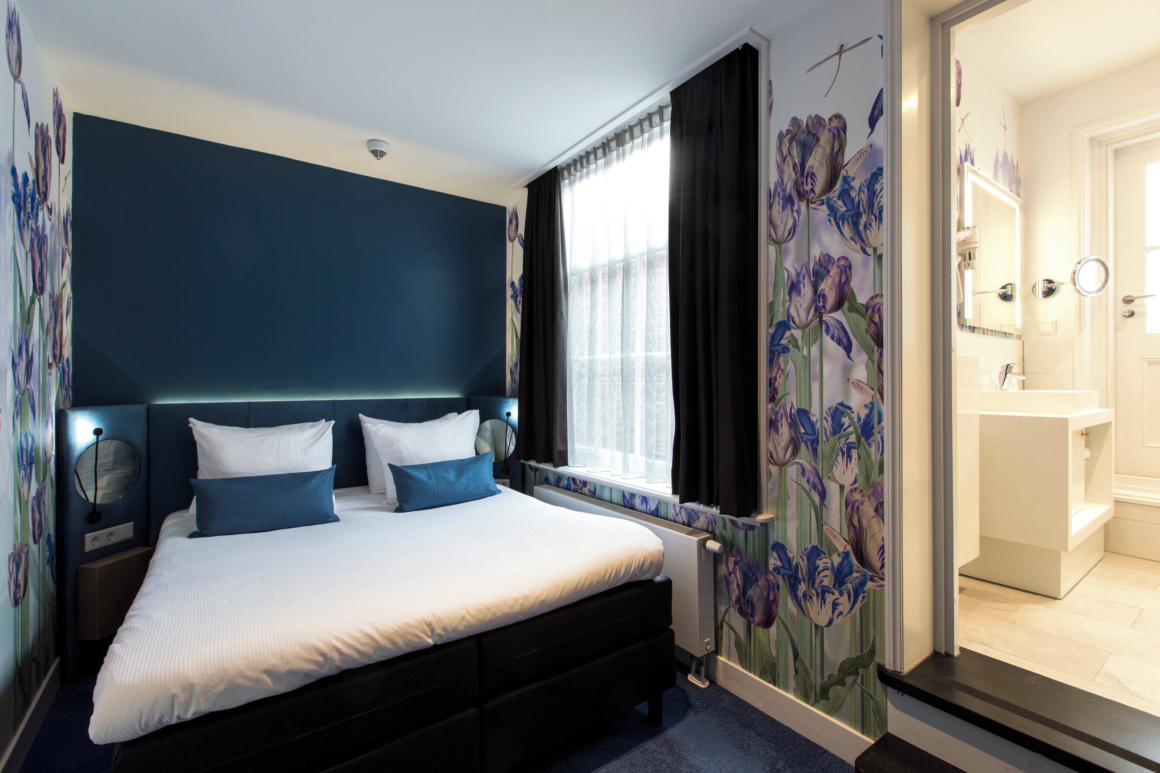 A bedroom with a large bed, a nightstand, a window with curtains, a bathroom with a mirror and sink.