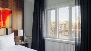 A hotel room with a bed, a nightstand, a lamp, a window with curtains, and a view of buildings and water. - Leonardo Eden Hotel Amsterdam City Center (Amsterdam)