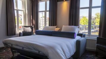 A hotel room with a large bed, a chair, a window with curtains, and a view of buildings and water.