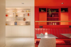 A modern kitchen with white cabinets and a red accent wall. There is a seating area with a table and a bench. - Leonardo Eden Hotel Amsterdam City Center (Amsterdam)