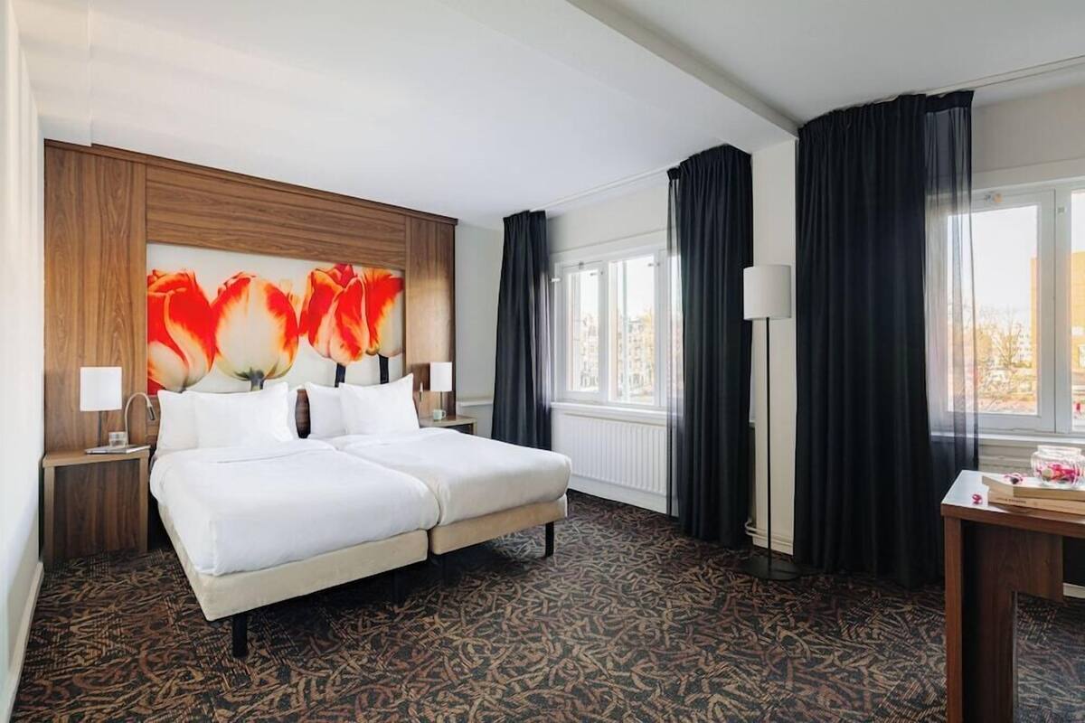 A hotel room with a large bed, a wooden headboard, a window with curtains, a nightstand, and a lamp.