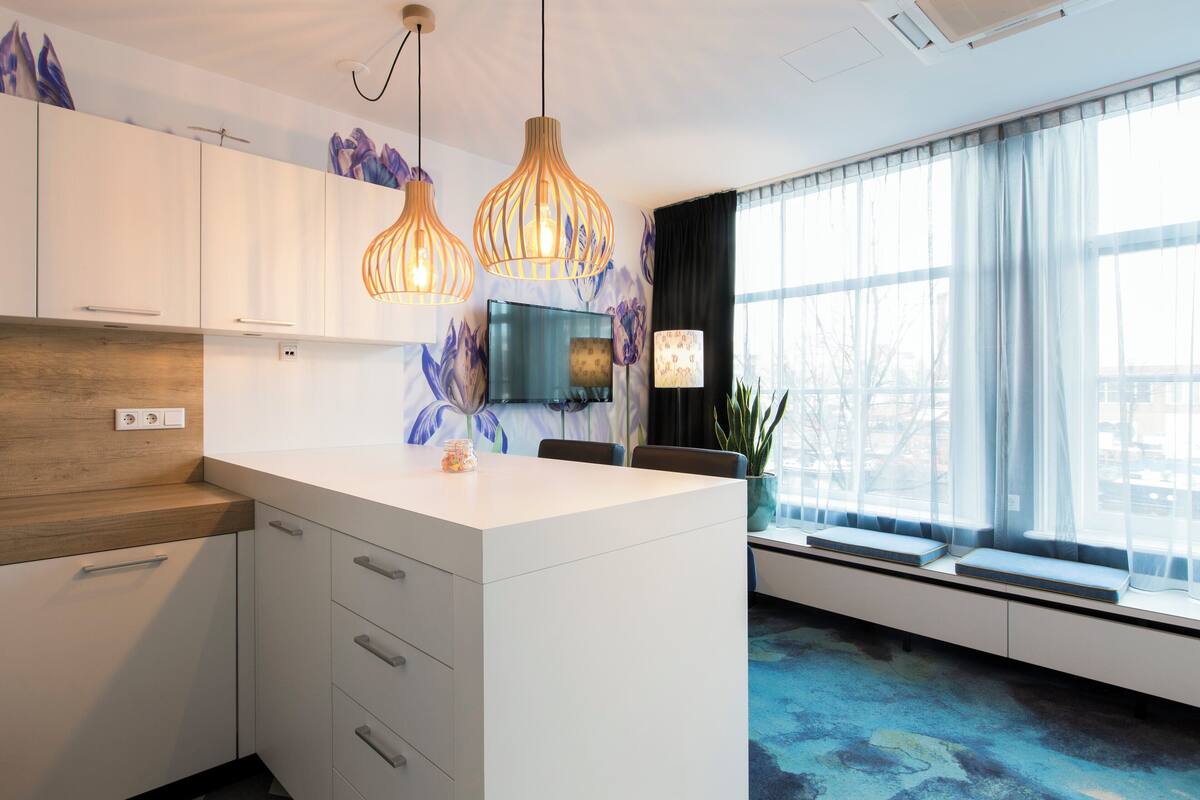 Una cocina moderna con armarios blancos, una isla central, luces colgantes, un televisor de pantalla plana y un amplio ventanal con cortinas traslúcidas.