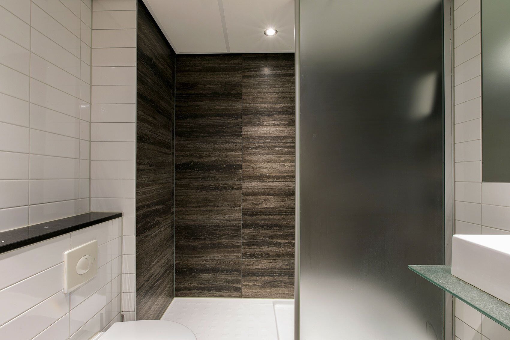Un baño moderno con azulejos blancos, una pared con textura de madera oscura, un box de ducha de vidrio y un inodoro blanco.