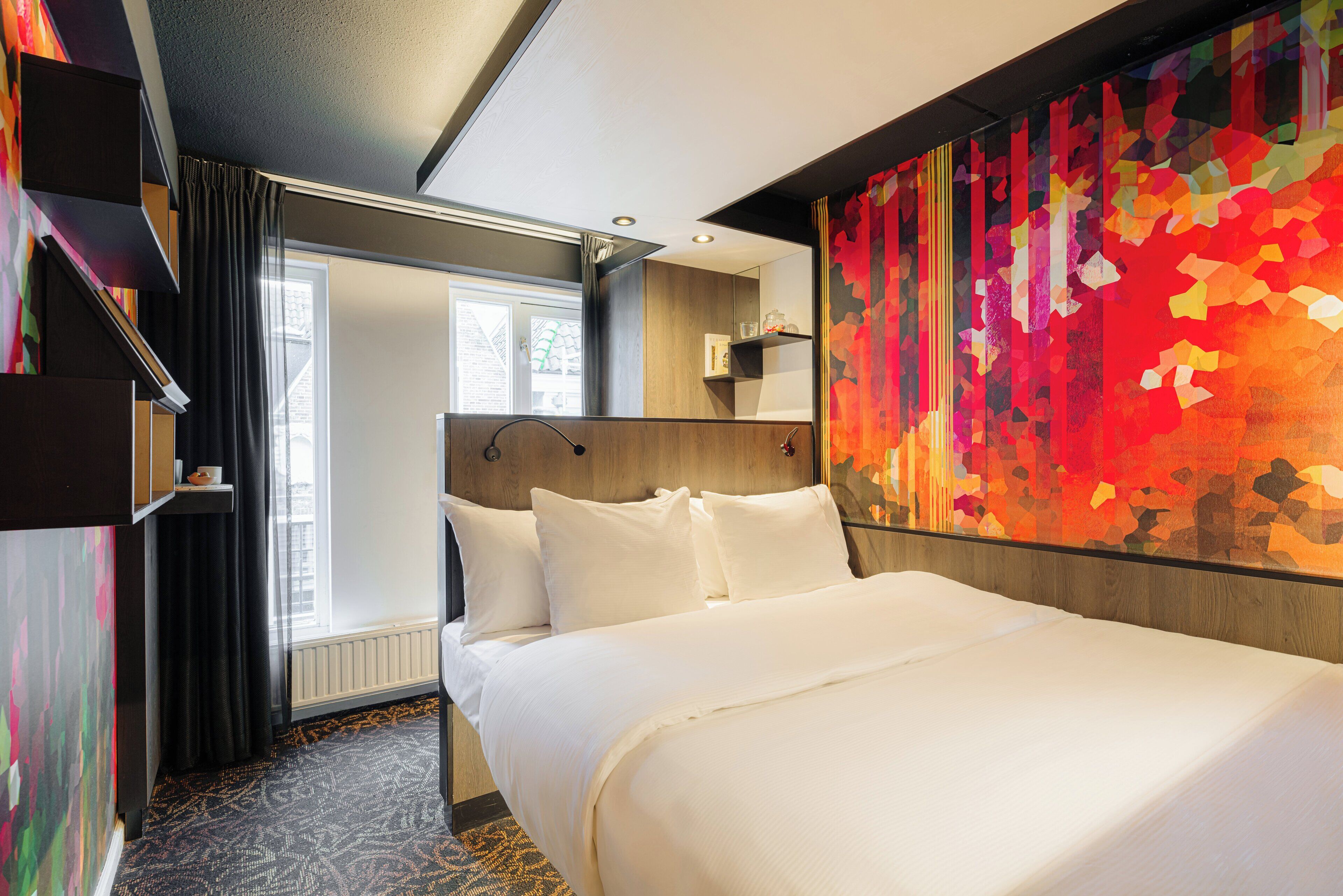 A hotel room with a large bed, a window with curtains, a colorful abstract wall, and a built-in shelf.