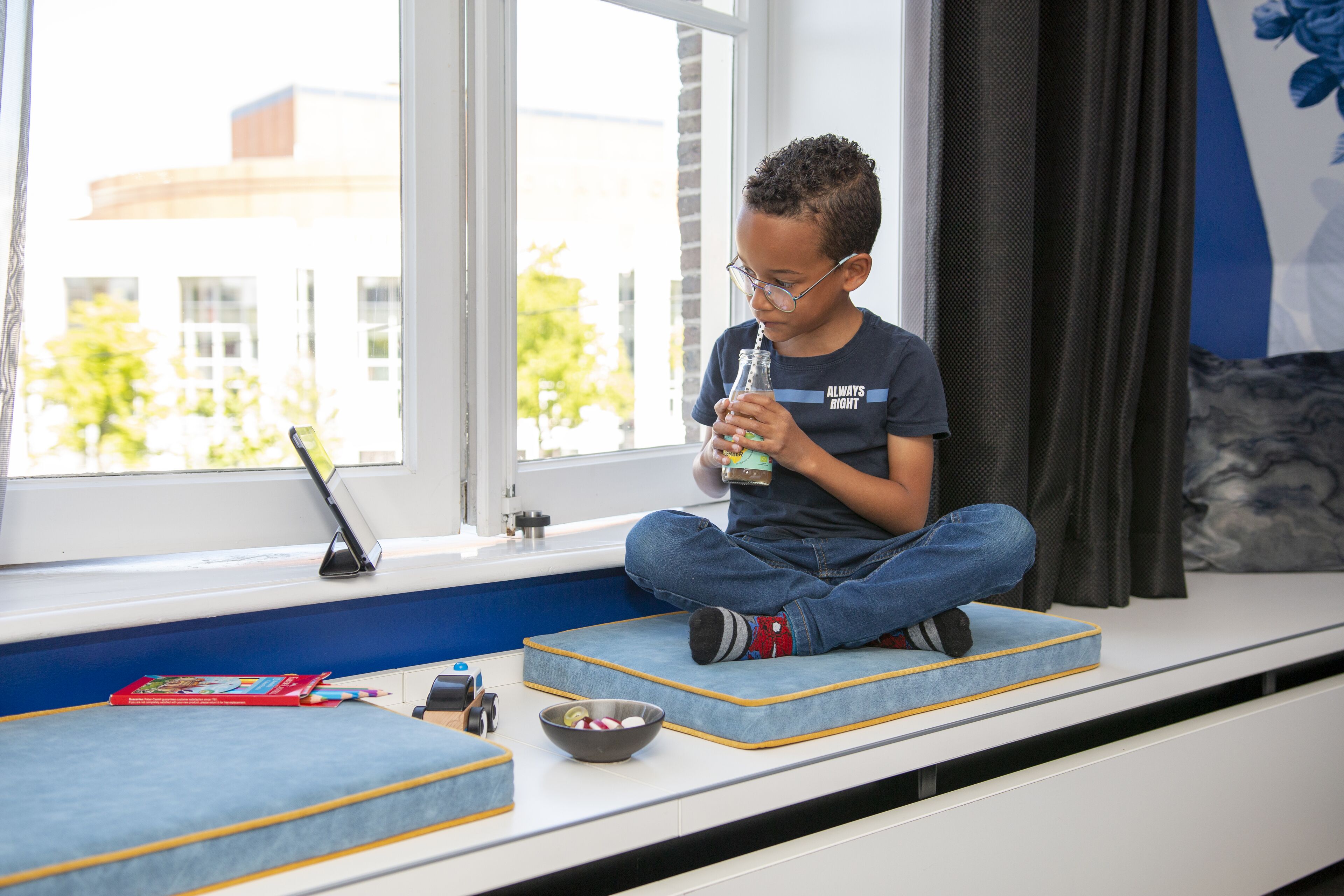 Een kind zit op een raamzitje met een laptop, een kom en een boek.