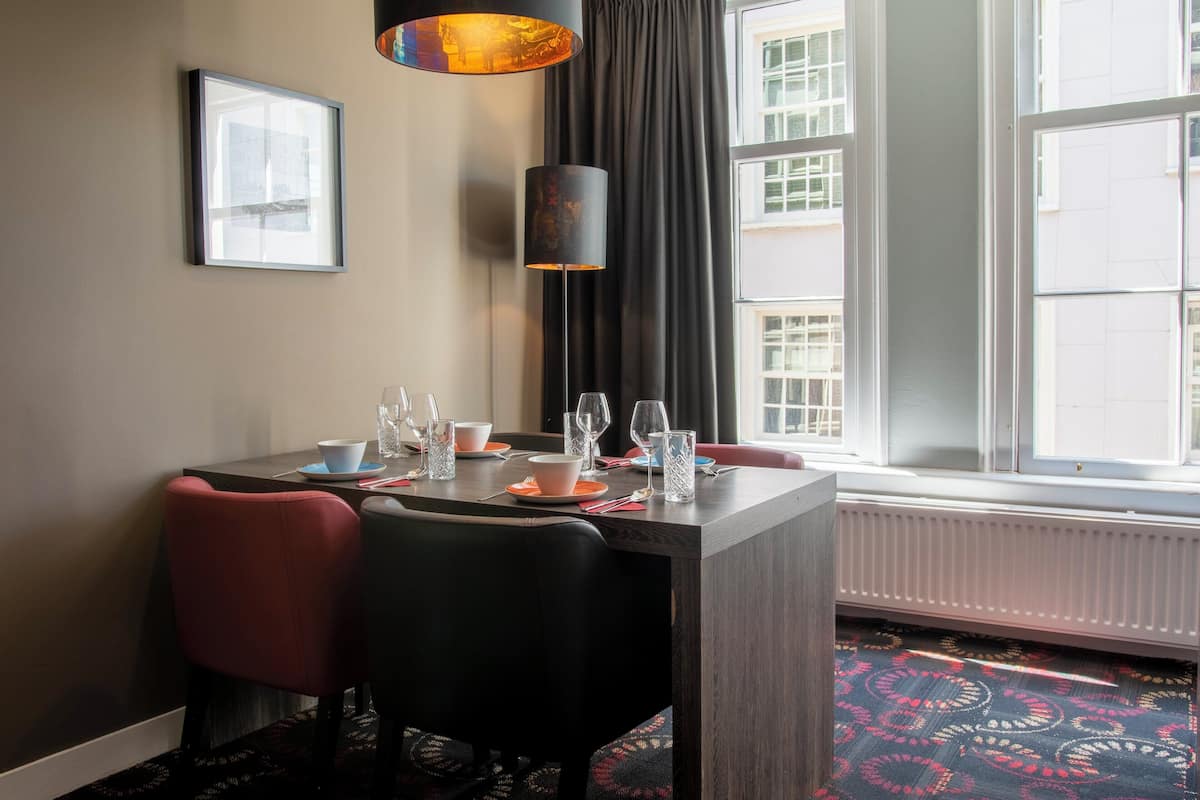 A dining area with a table set for two, a framed picture on the wall, a floor lamp, and a window with a view of buildings.