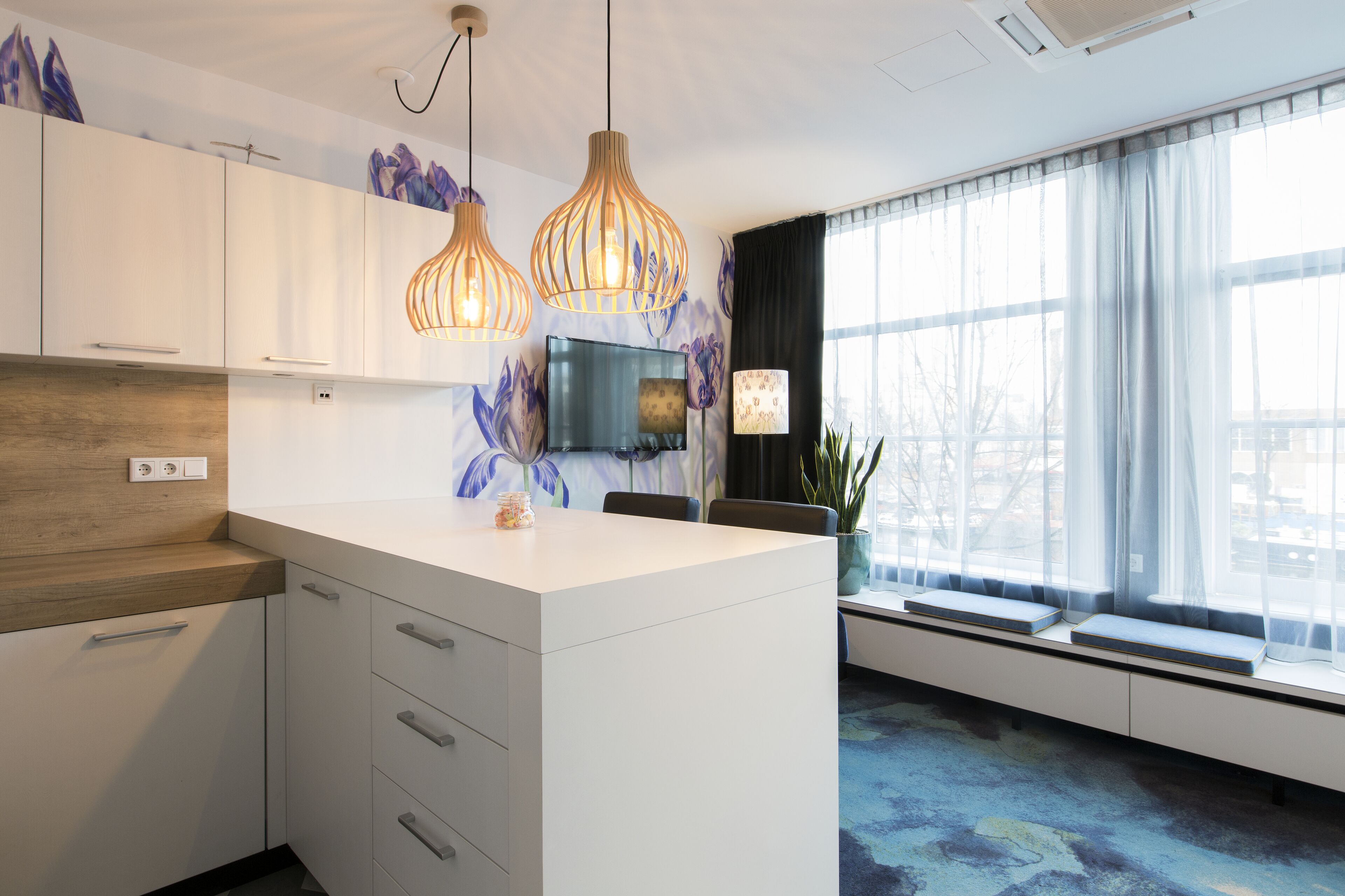 Una cocina moderna con armarios blancos, una isla central, luces colgantes y un amplio ventanal con cortinas translúcidas.