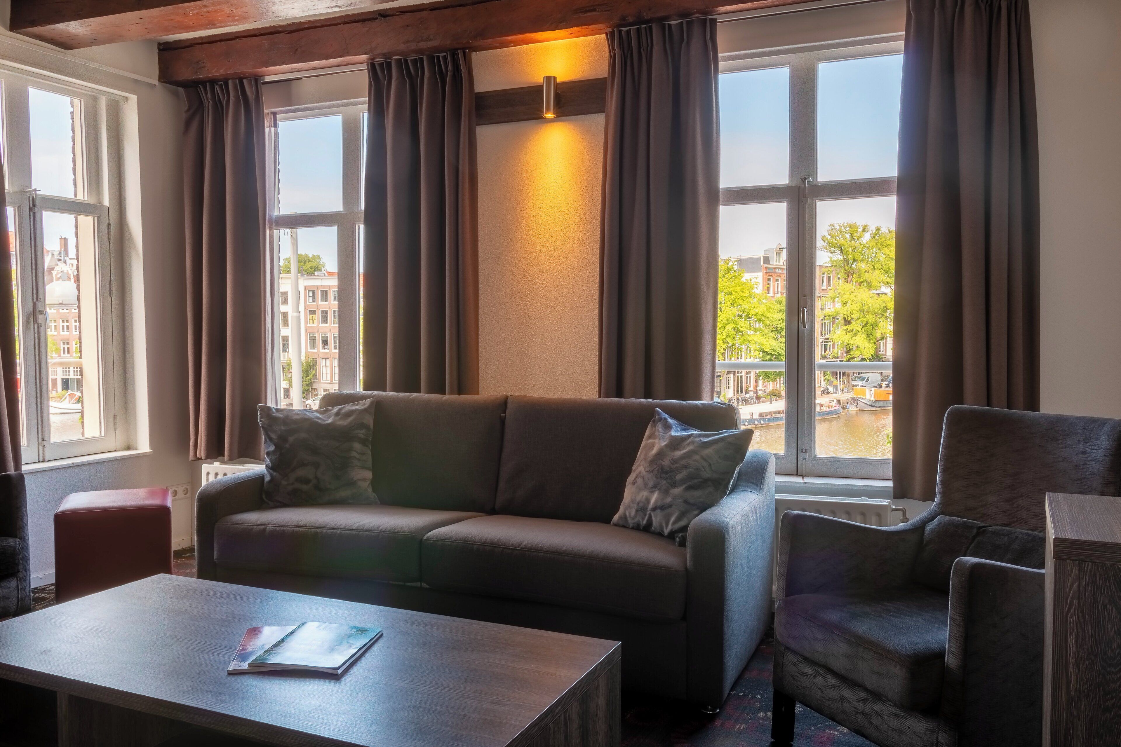 Een moderne woonkamer met een bank, fauteuil en salontafel. Grote ramen bieden uitzicht op gebouwen en een gracht.