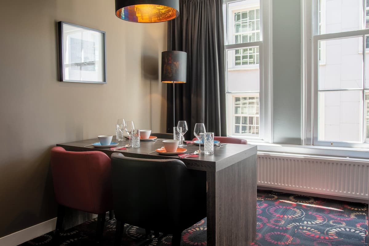 Un comedor con una mesa dispuesta para dos, un cuadro en la pared, una lámpara de pie y un ventanal con vistas a edificios.
