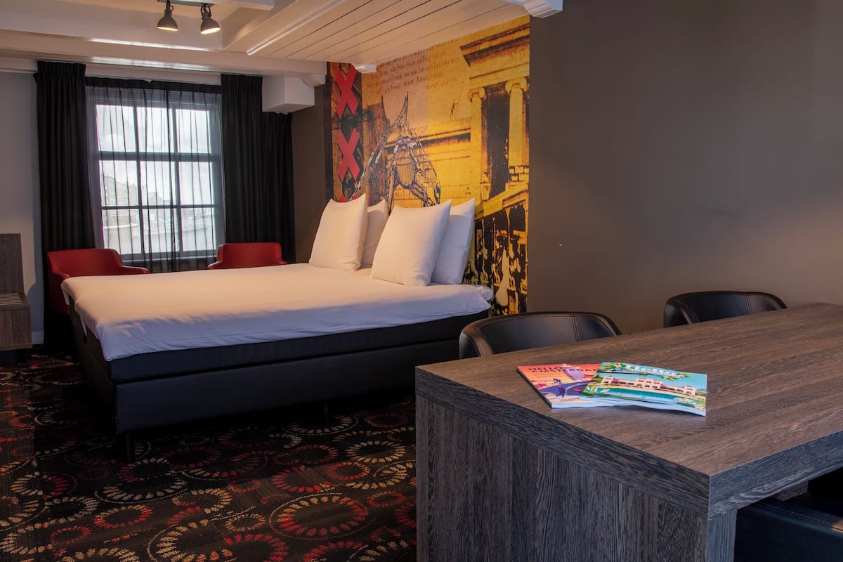 A hotel room with a large bed, a desk, and a chair. There is a window with curtains, a wall with a mural, and a patterned carpet.