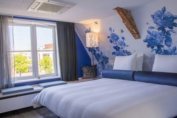A modern hotel room with a large bed, a window with a view of buildings, a blue wall with floral wallpaper, and a nightstand with a lamp. - Leonardo Eden Hotel Amsterdam City Center (Amsterdam)