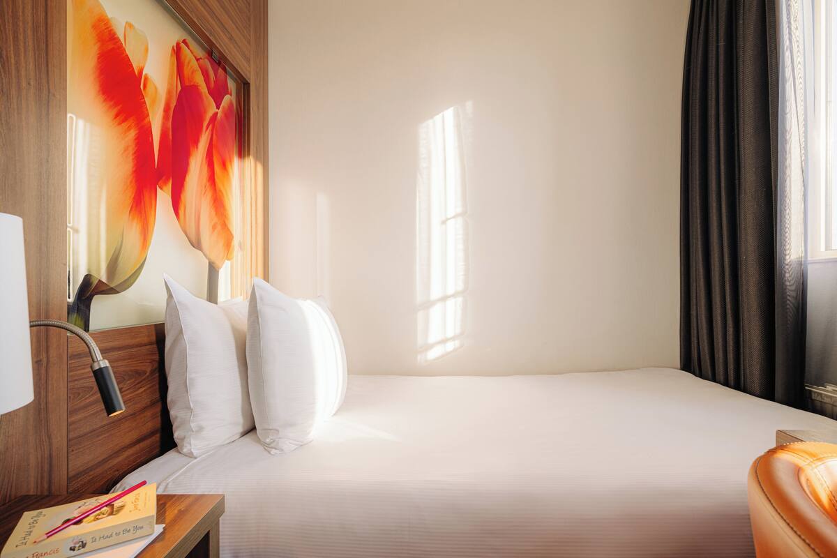 Habitación de hotel con cama, mesita de noche, lámpara, ventana con cortinas y un cuadro grande con motivos florales en la pared.