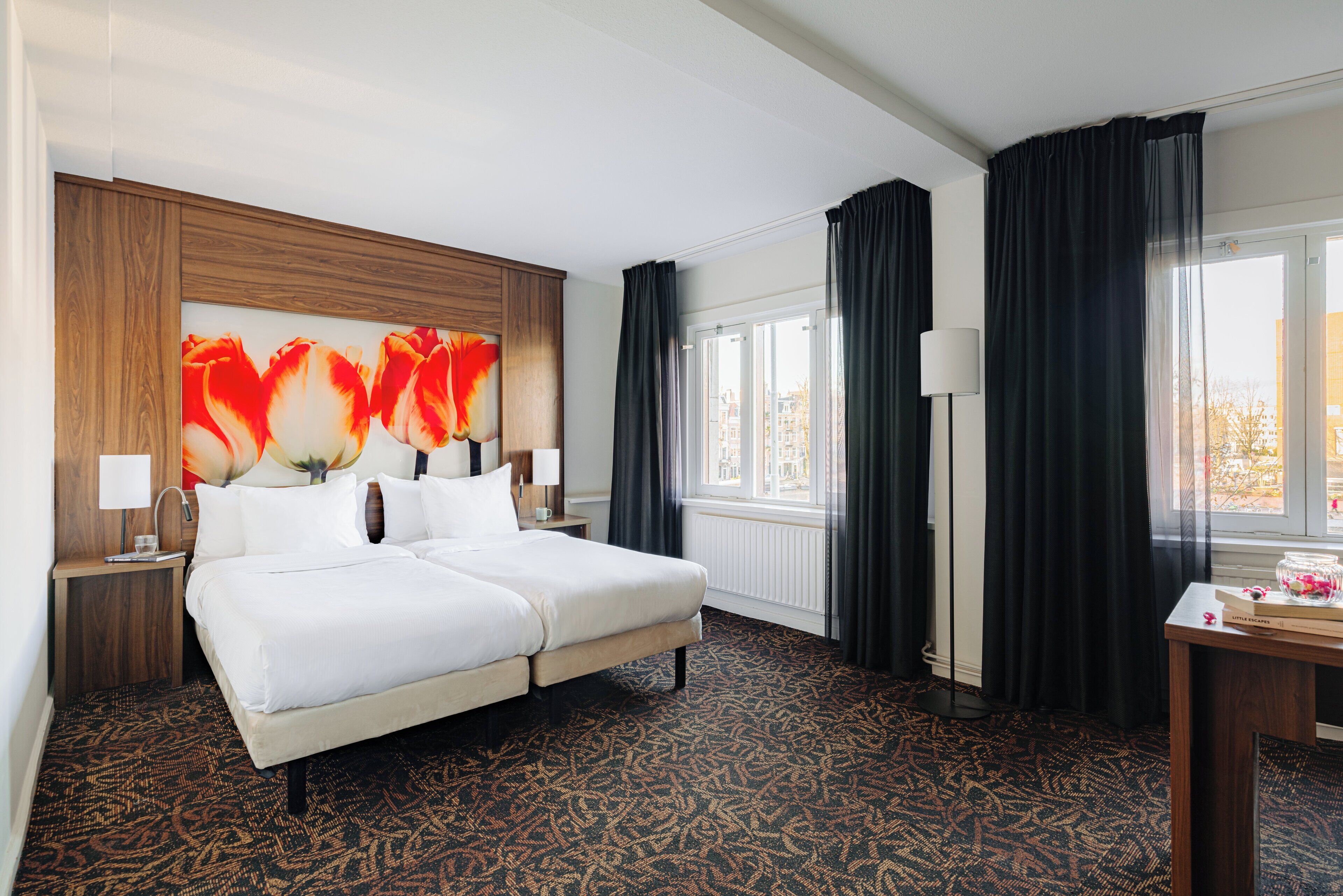A hotel room with a large bed, a wooden headboard, a large window with curtains, and a patterned carpet.