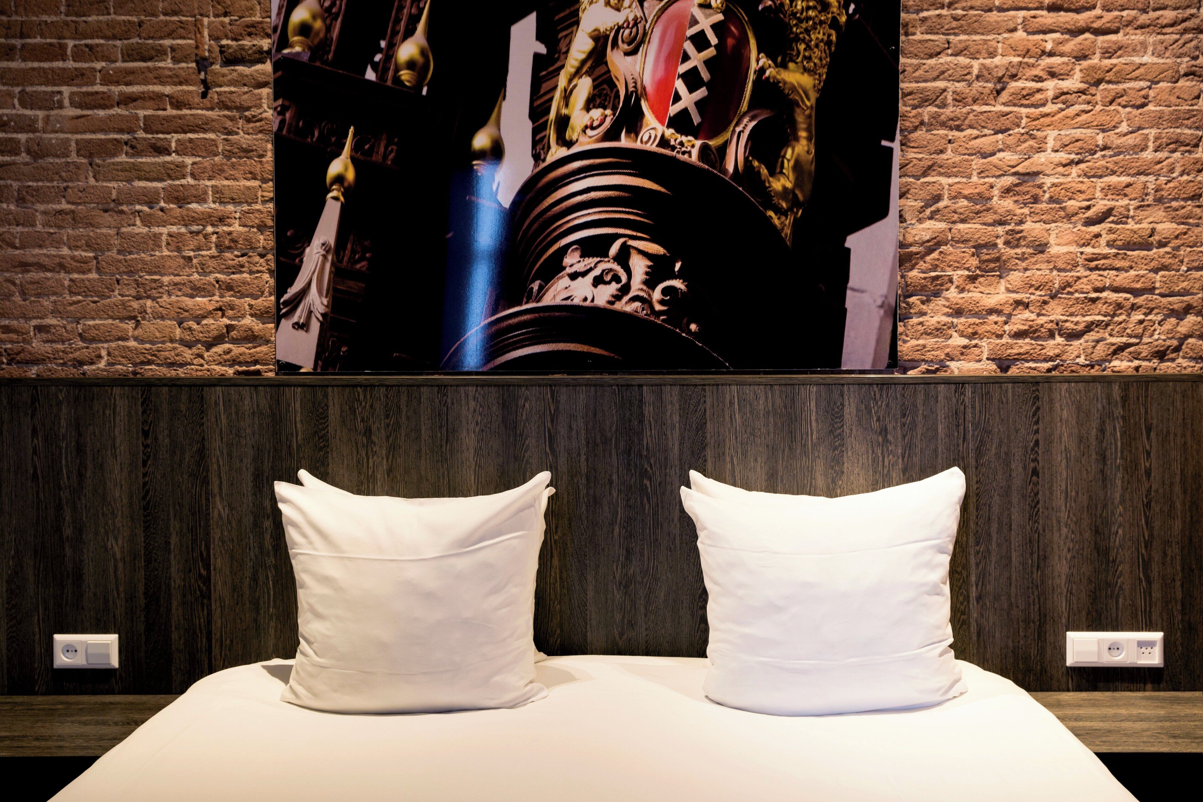 Een hotelkamer met een bakstenen muur, een groot decoratief schilderij en twee witte kussens op het bed.