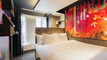 Une chambre d’hôtel avec un grand lit, une fenêtre avec des rideaux, un mur décoré d’une œuvre abstraite aux couleurs vives et une étagère intégrée.