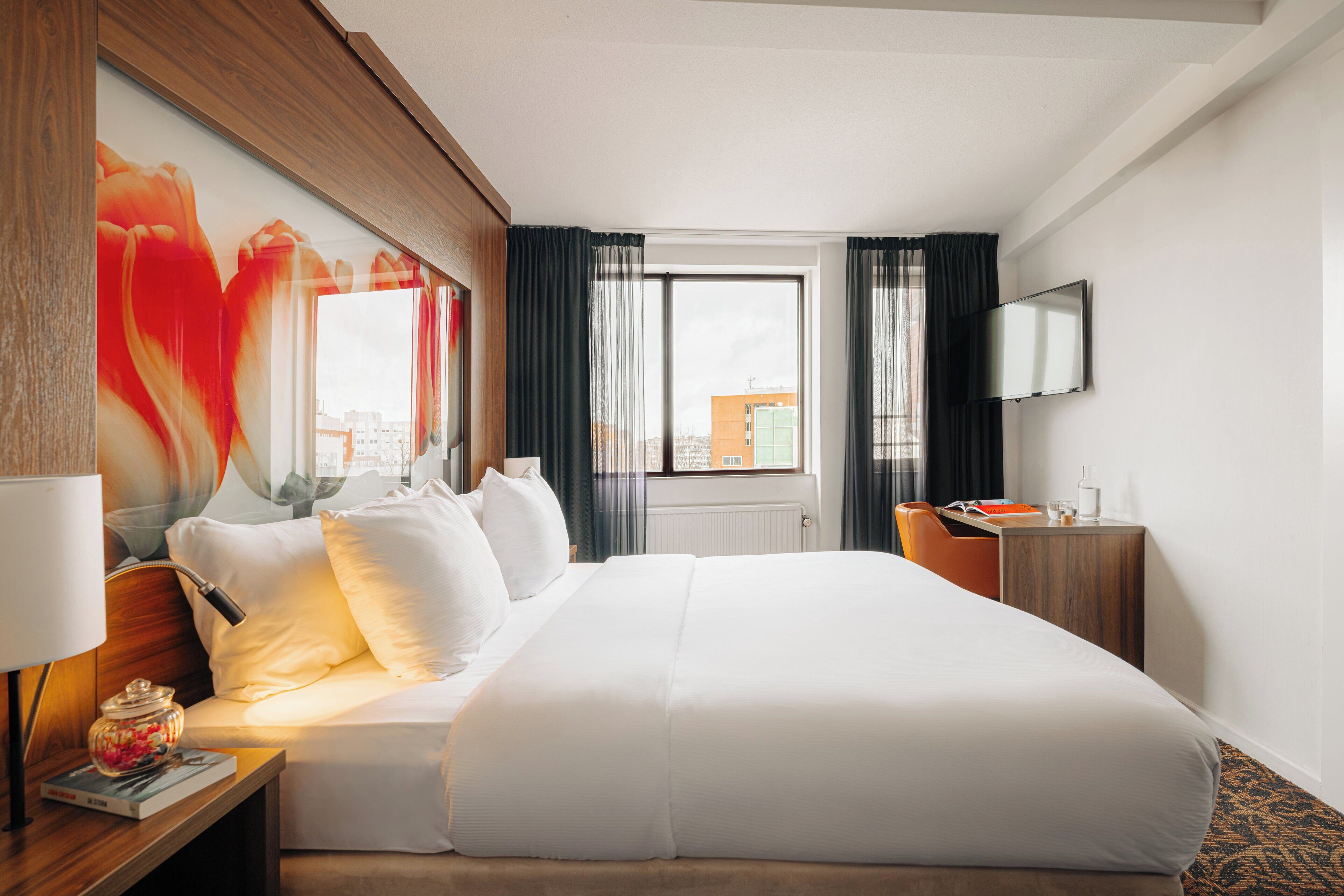 Une chambre d’hôtel avec un grand lit, un bureau avec une lampe, une télévision et une fenêtre avec des rideaux.