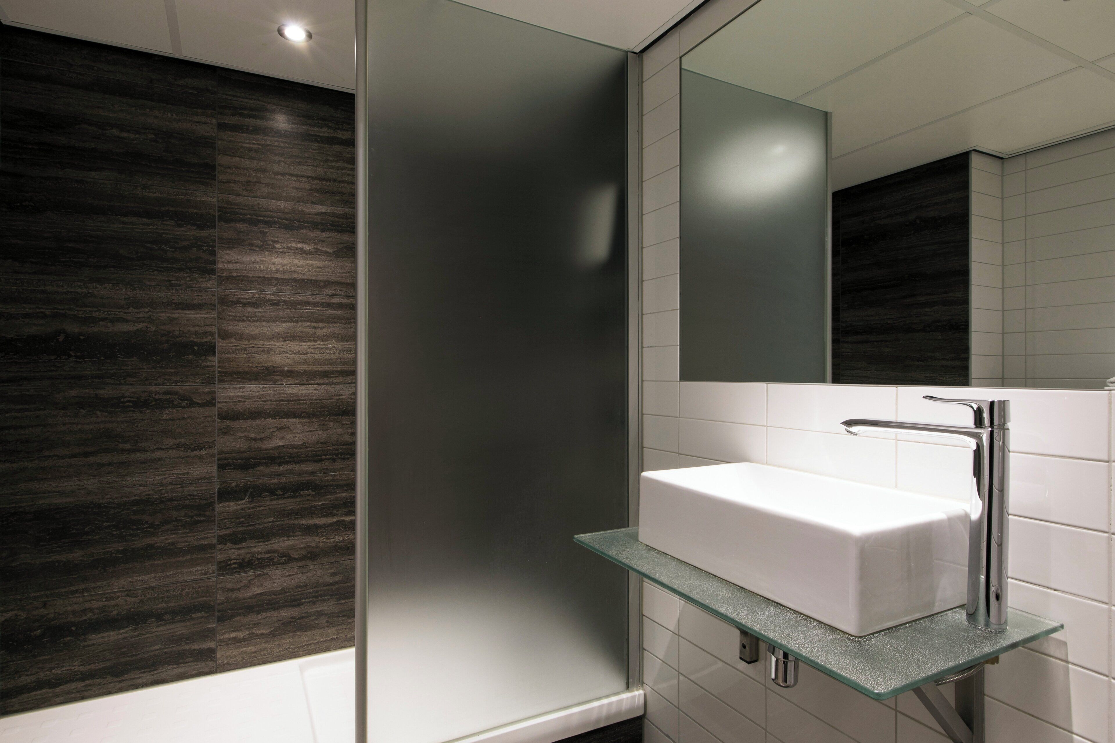 A modern bathroom with a glass shelf holding a white sink, a mirror above it, and dark wood paneling on the walls.