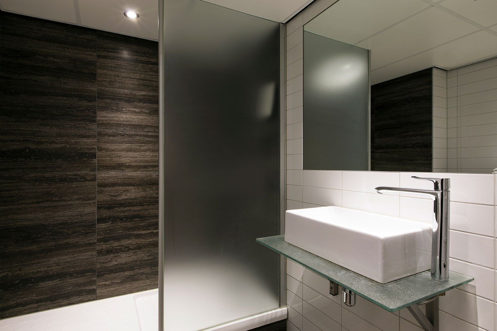 Un baño moderno con una repisa de vidrio que sostiene un lavamanos blanco, paneles de madera oscura en las paredes y un espejo grande sobre el lavamanos.