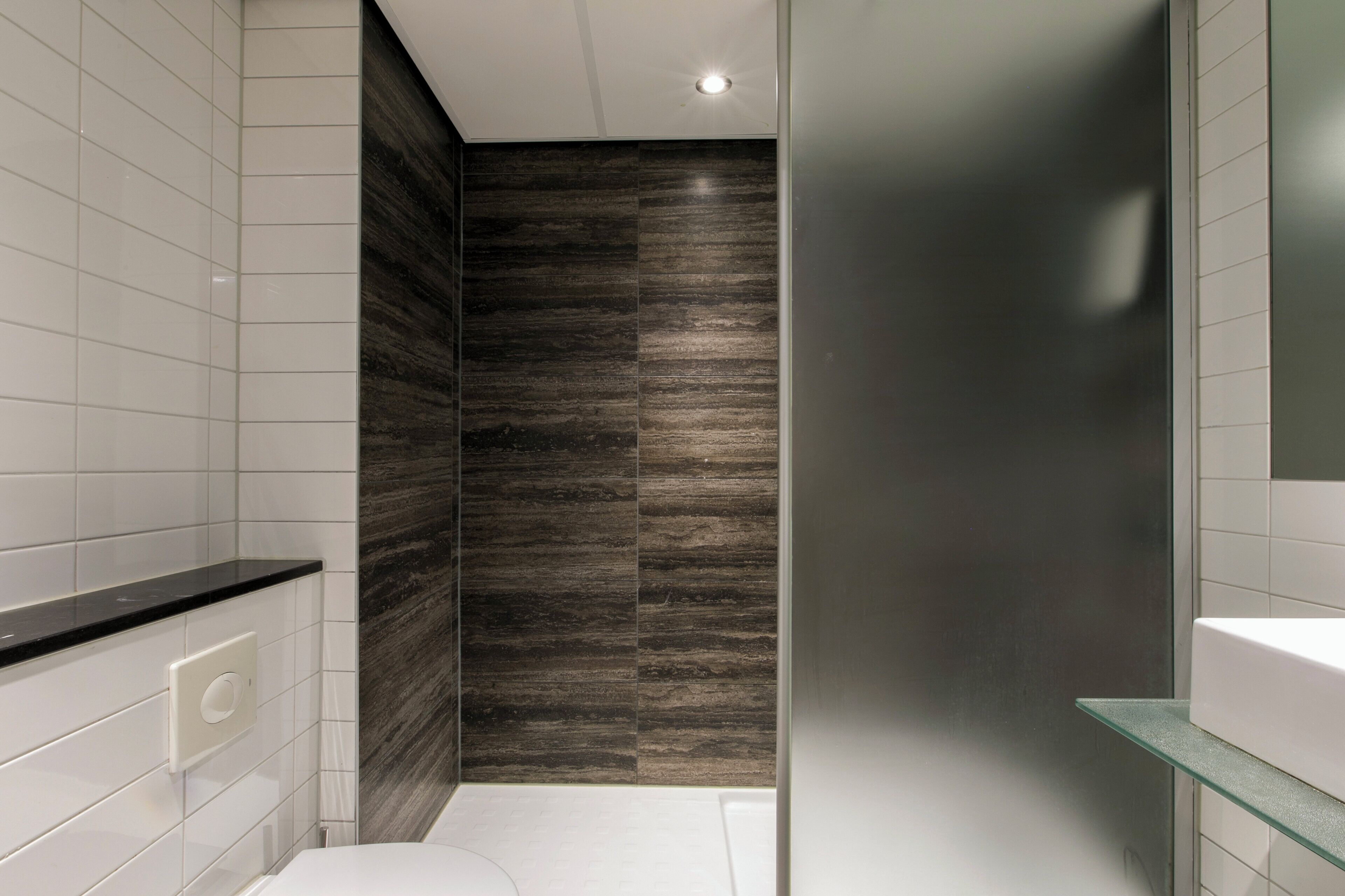 A modern bathroom with white tiles, a dark wood panel wall, a glass shower enclosure, and a white sink with a glass shelf.