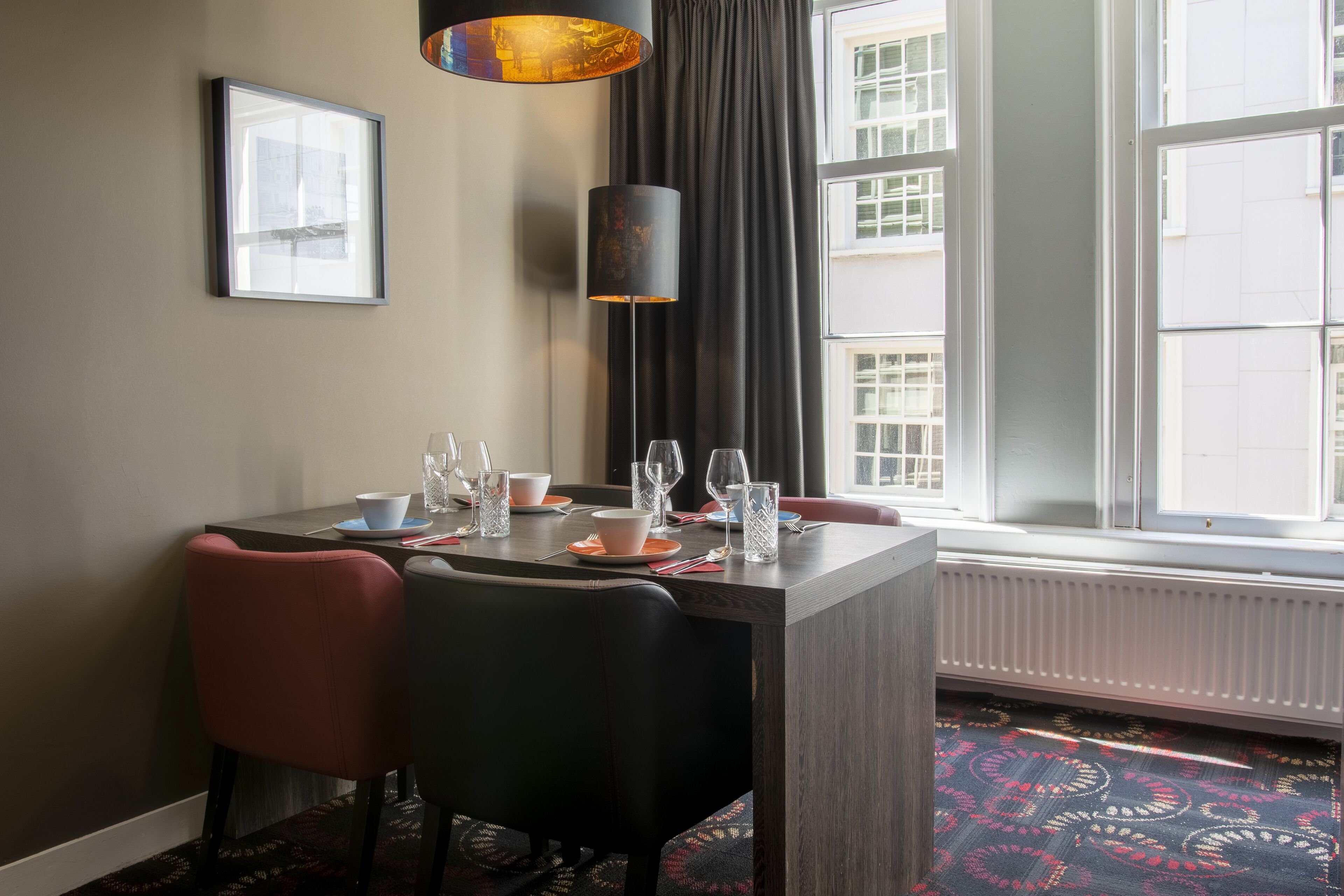 Un comedor con una mesa dispuesta para dos personas, una silla negra, una silla roja, una lámpara de pie y un ventanal con vistas a edificios.