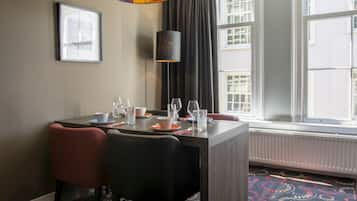 A dining area with a table set for two, a black chair, a red chair, a floor lamp, and a window with a view of buildings.