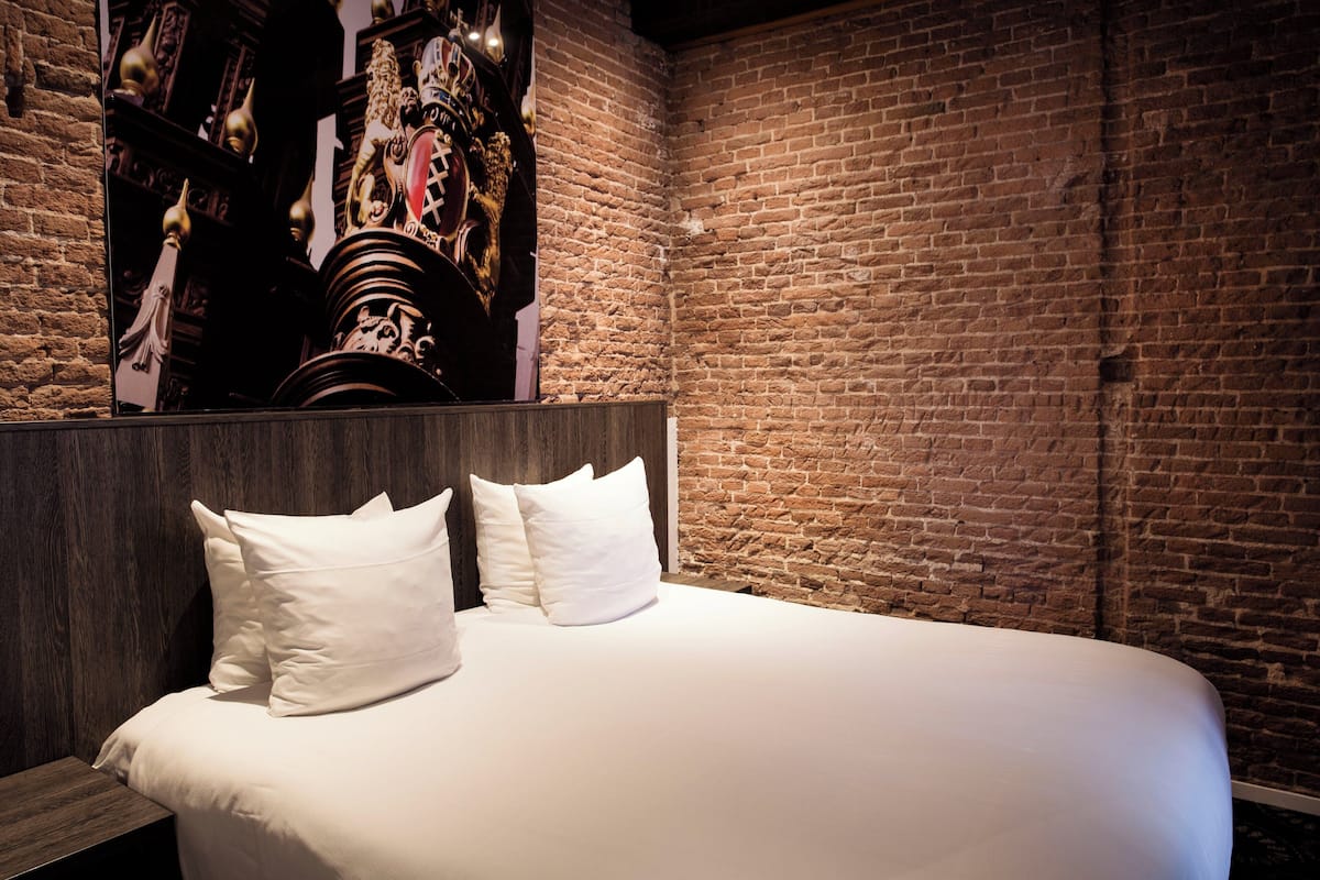 A neatly made bed with white linens and pillows, a brick wall, and a wooden headboard.