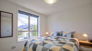 Une chambre avec une grande fenêtre, un lit recouvert d’une couvre-lit à motifs géométriques, une table de chevet avec une lampe et un luminaire fixé au mur.