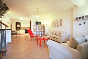 A spacious living area with a sofa, red chair, and a dining table. A kitchen is visible in the background with a TV on the counter.