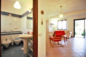 A bathroom with a sink, bidet, and toilet. There is a chair in a living area with a view of the outdoors.