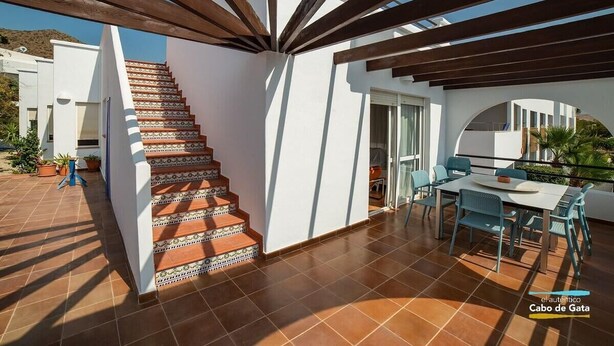 Un patio moderno al aire libre con una pérgola de madera, una escalera con baldosas estampadas y una zona de comedor con sillas y mesa azules.