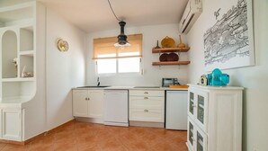 Una cocina con armarios blancos, un ventanal con persianas, una lámpara colgante y un aire acondicionado instalado en la pared.