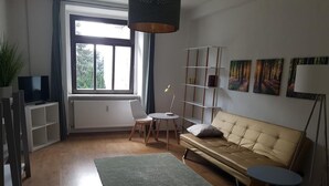Ein Wohnzimmer mit einer Couch, einem kleinen runden Tisch, einer Stehlampe, einem Bücherregal und einem großen Fenster.