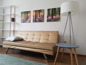 A modern living room with a beige sofa, a small round table, and a floor lamp. There are three framed pictures on the wall depicting forest scenes. - Dresdner-Tag Linda (Dresden)