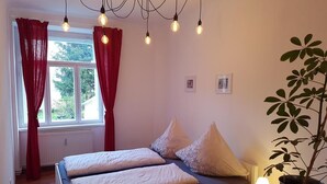 Ein Schlafzimmer mit zwei Betten, roten Vorhängen, einem Blumentopf und einem Fenster mit Blick auf Grünflächen.